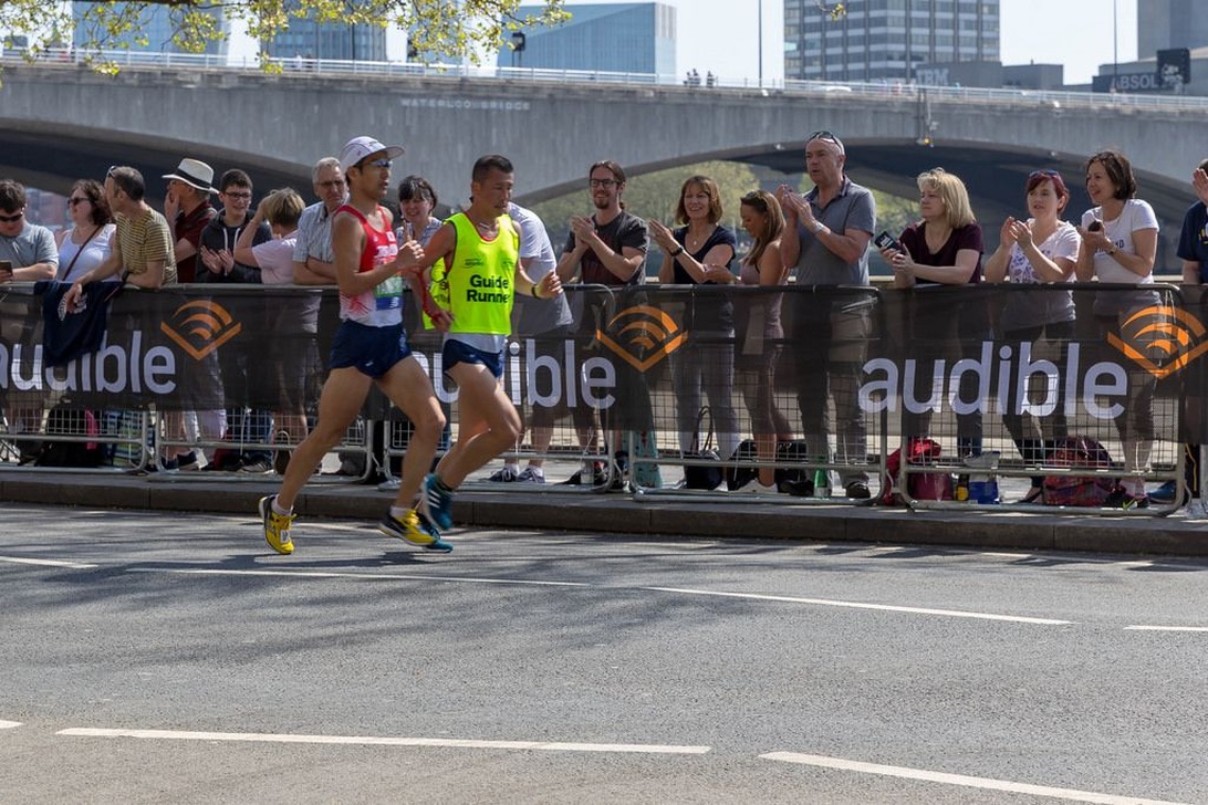 Marathonläufer in einer städtischen Umgebung tragen leichte Rennausrüstung. Zwei Läufer im Vordergrund tragen Führwesten, während Zuschauer an Absperrungen jubeln. Die helle städtische Umgebung und die minimale Kleidung der Läufer deuten auf warme Wetterbedingungen hin, die für Sommerläufe typisch sind.